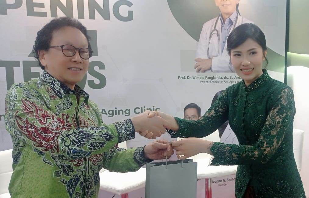 Four people seated at a grand opening event, one man is speaking into a microphone while others listen; backdrop shows 'Grand Opening Steros' with medical imagery.