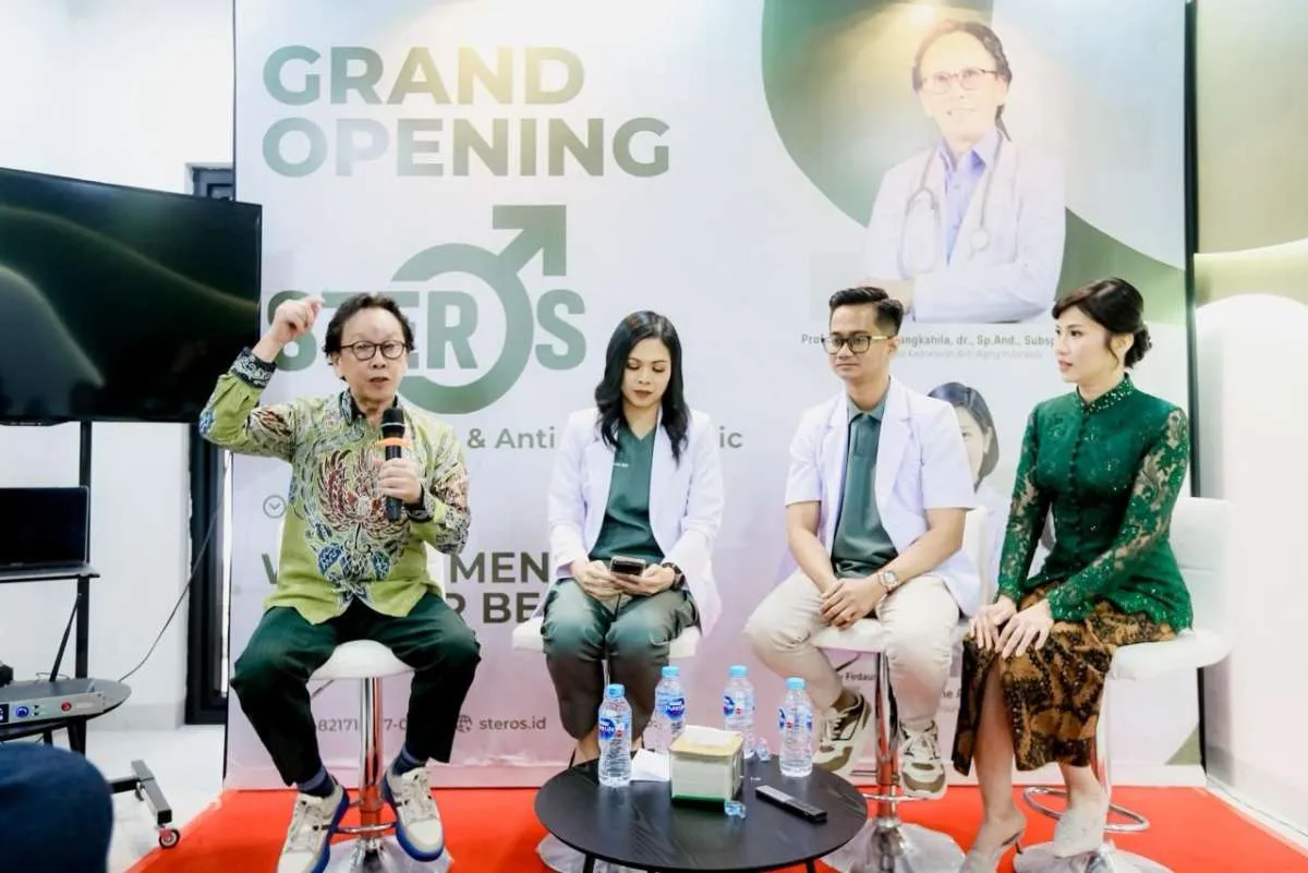 Four people seated at a grand opening event, one man is speaking into a microphone while others listen; backdrop shows 'Grand Opening Steros' with medical imagery.