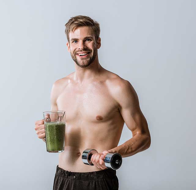 Shirtless young man smiling while holding a green smoothie in one hand and a dumbbell in the other.