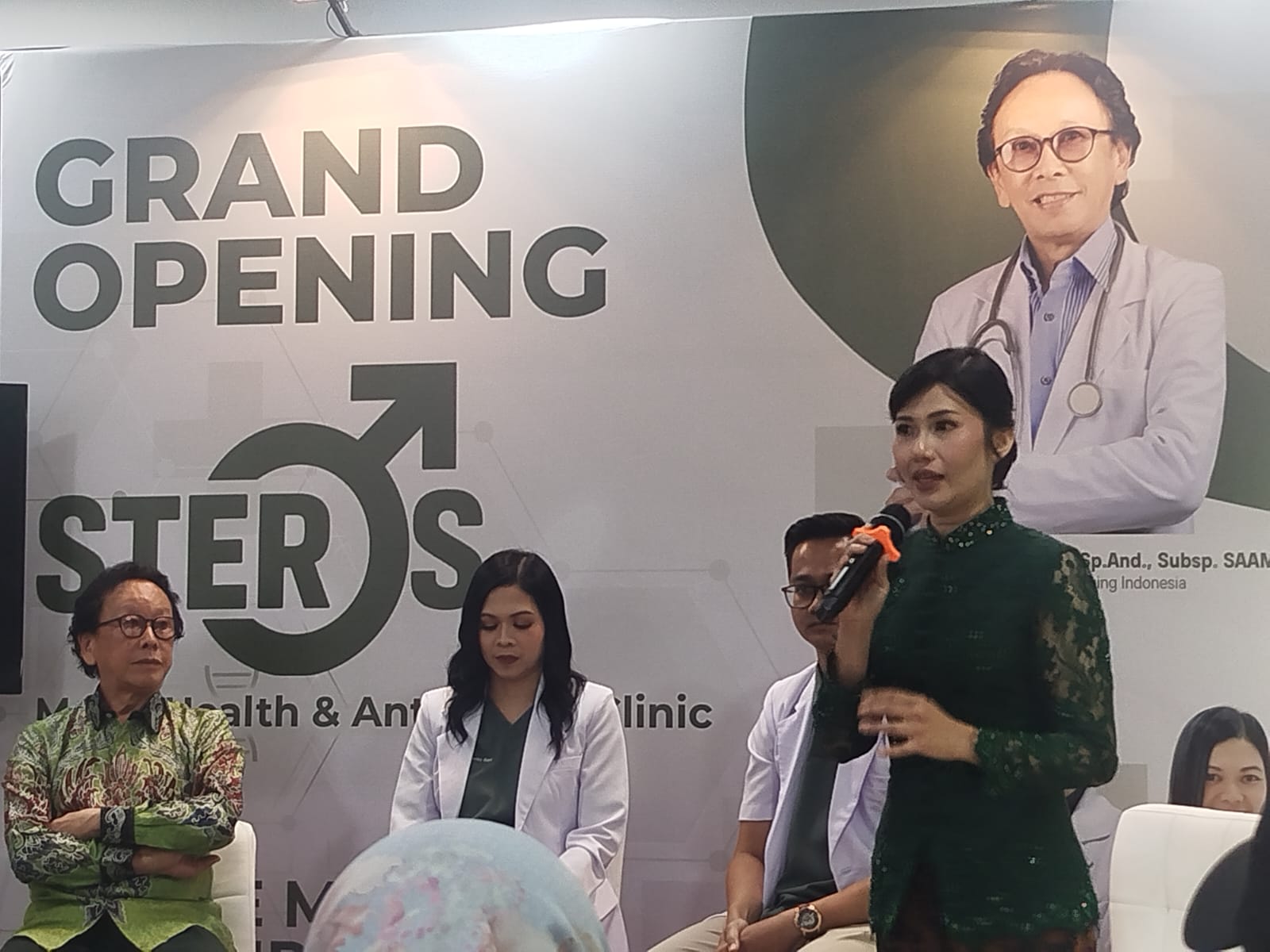 Four people seated at a grand opening event, one man is speaking into a microphone while others listen; backdrop shows 'Grand Opening Steros' with medical imagery.